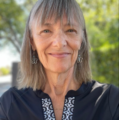 Laurie Mook headshot - fair-skinned woman, smiling, gray hair, navy shirt, earrings