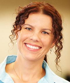 Rebekka Goodman headshot - fair-skinned woman, smiling, brown hair, light blue collared shirt, necklace, earrings